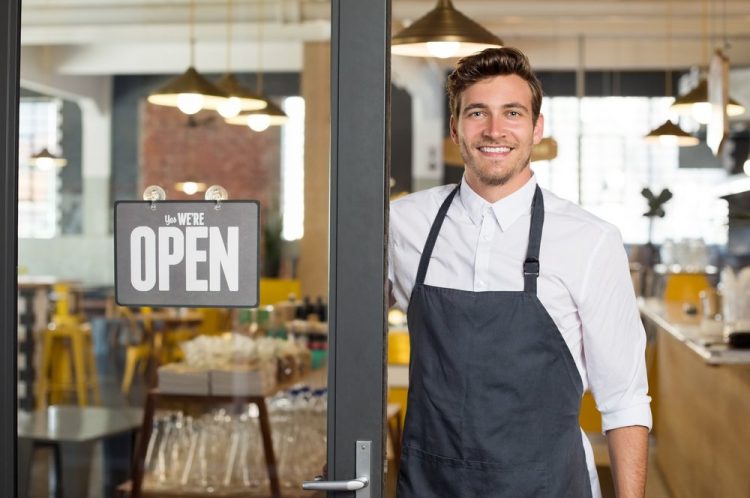 Smart young man in an apron standing by the door to a café.